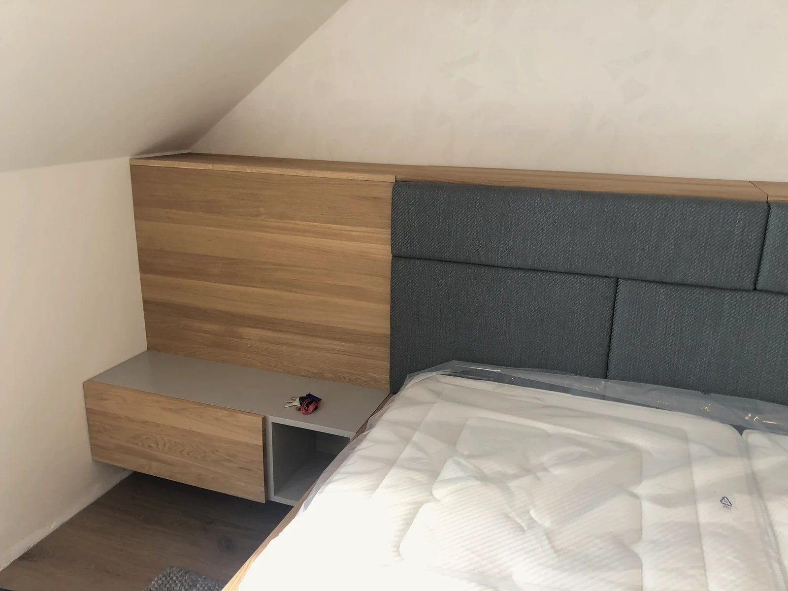 Detail čela postele z dubového masivu s šedými čalouněnými panely a závěsný noční stolek s dubovou šuplíkovanou částí a šedou deskou